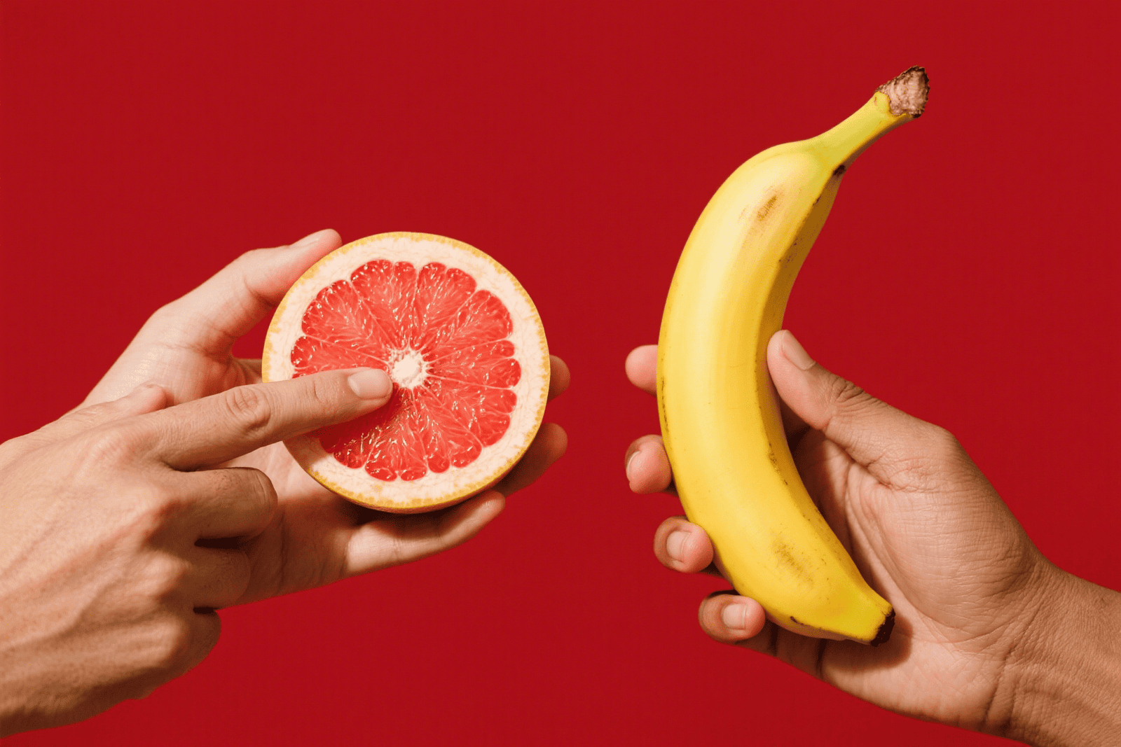 Two hands holding half a grapefruit and a banana against a red background
